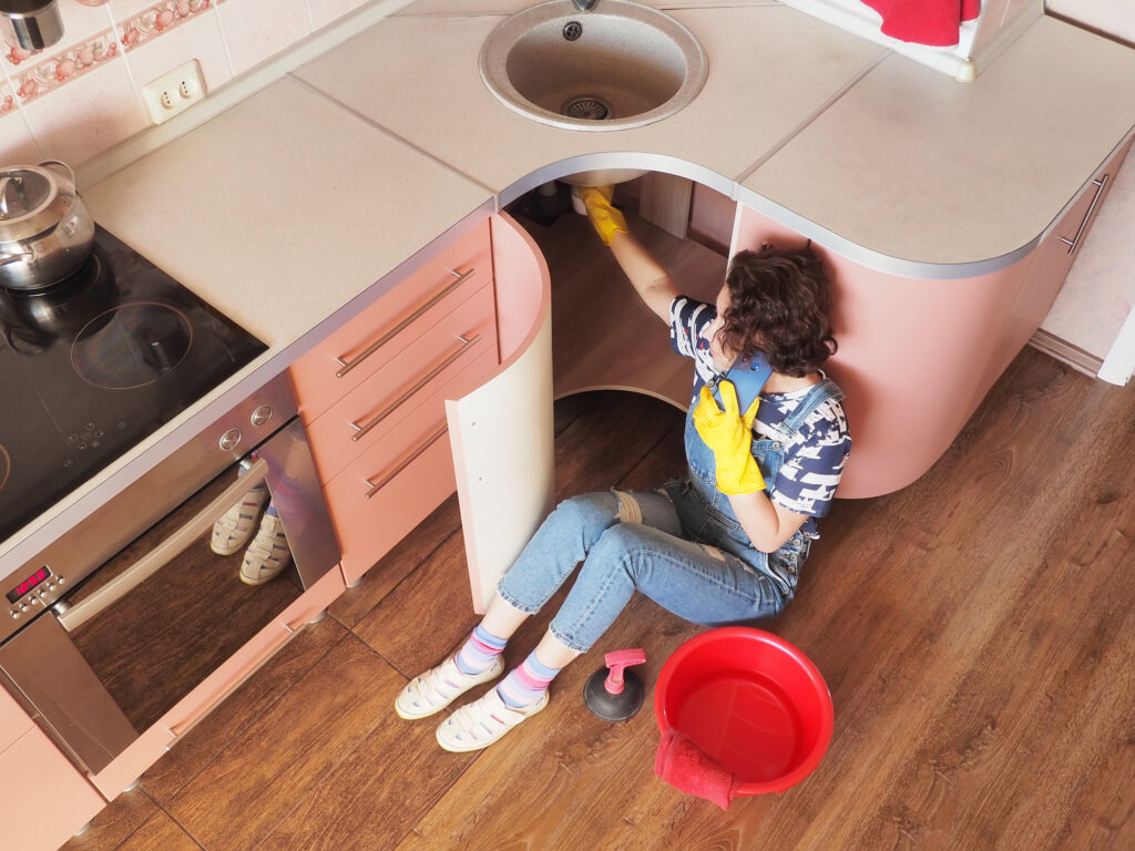 A young woman sitting on the floor trying to stop a kitchen leak while she speaks on the phone with a plumber.