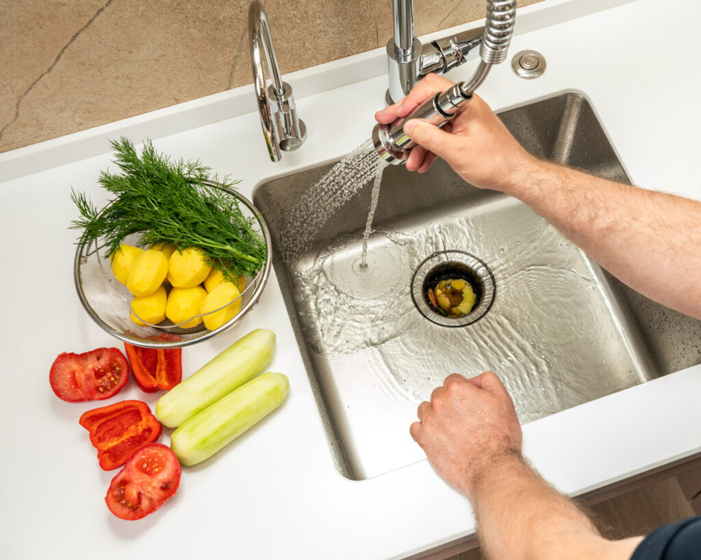 A person washing a food item down the garbage disposal with various vegetables on the counter next to it in Waldorf.